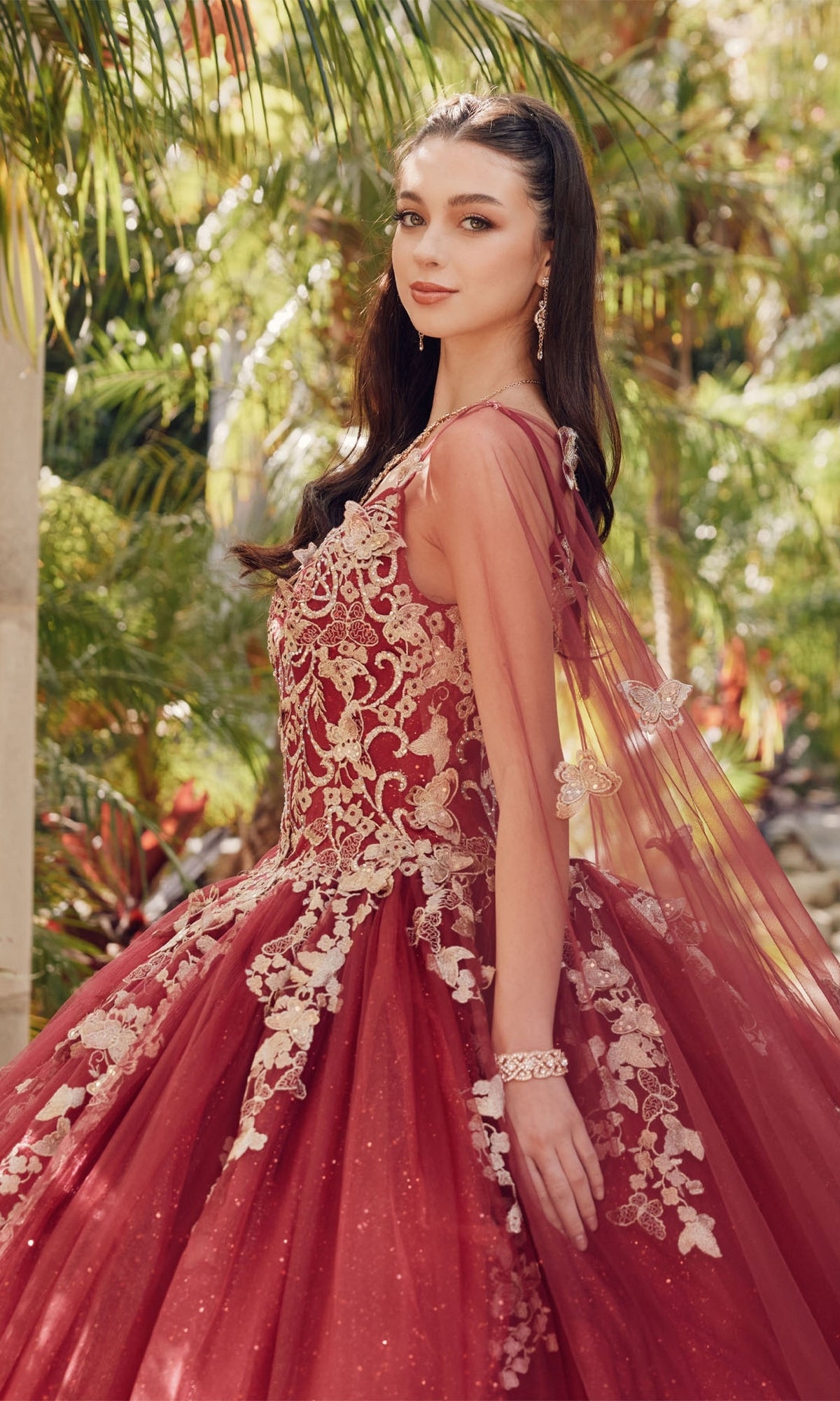 Wearing the Juliet 1442 Quinceañera Dress with a sheer cape, a woman stands outdoors among green plants, glancing over her shoulder.